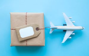 Flat lay of wrapped package with blank tag and airplane model on pastel blue background. Shipping logistics transport minimal creative concept.