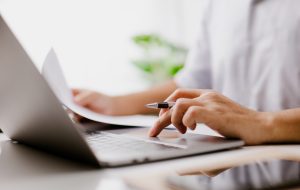 Businessman hand is on the trackpad, typing on a laptop keyboard, while reading a business document. In a modern office, a corporate man, a lawyer, works. Concept of business and technology.