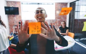 Smiling businesswoman sticking adhesive notes to a glass wall with her colleagues in the background. Happy young businesswoman sharing her ideas with her team in a modern office.