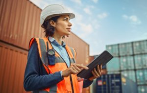 Smiling Portrait of a Beautiful Latin Female Industrial Engineer in White Hard Hat, High-Visibility Vest Working on Tablet Computer. Inspector or Safety Supervisor in Container Terminal.