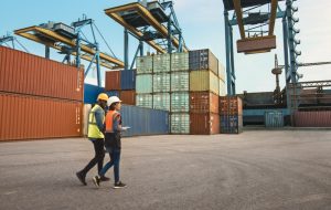 Multiethnic Female Industrial Engineer with Tablet and Black African American Male Supervisor in Hard Hats and Safety Vests Walk in Container Terminal. Colleagues Talk About Logistics Operations.