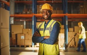 Handsome and Happy Professional Worker Wearing Safety Vest and Hard Hat Smiling with Crossed Arms on Camera. In the Background Big Warehouse with Shelves full of Delivery Goods. Medium Portrait