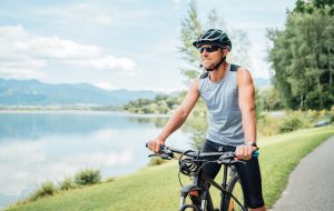 Portrait of a sincerely smiling man dressed in cycling clothes, helmet and sunglasses riding a bicycle on the asphalt out-of-town bicycle path along a mount lake. Active sporty people concept image.