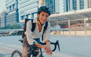 Asian businesswoman go to work at office stand and smiling wear backpack look at camera with bicycle on street around building on a city. Bike commuting, Commute on bike, Business commuter concept.