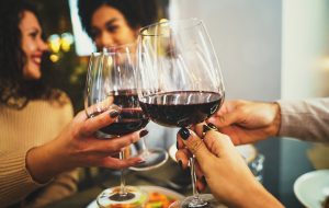 Interracial happy friends clinking red wine glasses at restaurant - group of young people having fun toasting - girlfriends watching in each other eyes on background - focus on the wineglasses.