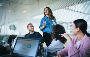 Businesswoman walking in office boardroom and sharing a strategy with team sitting at table. Female manager explaining new business plan with coworkers sitting at conference table in office.