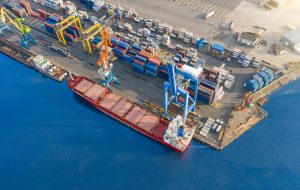 Aerial top view huge cargo ship moored at the pier at the port, loading goods, ore mineral bulk cargo in railway cars, concrete and other solid raw materials